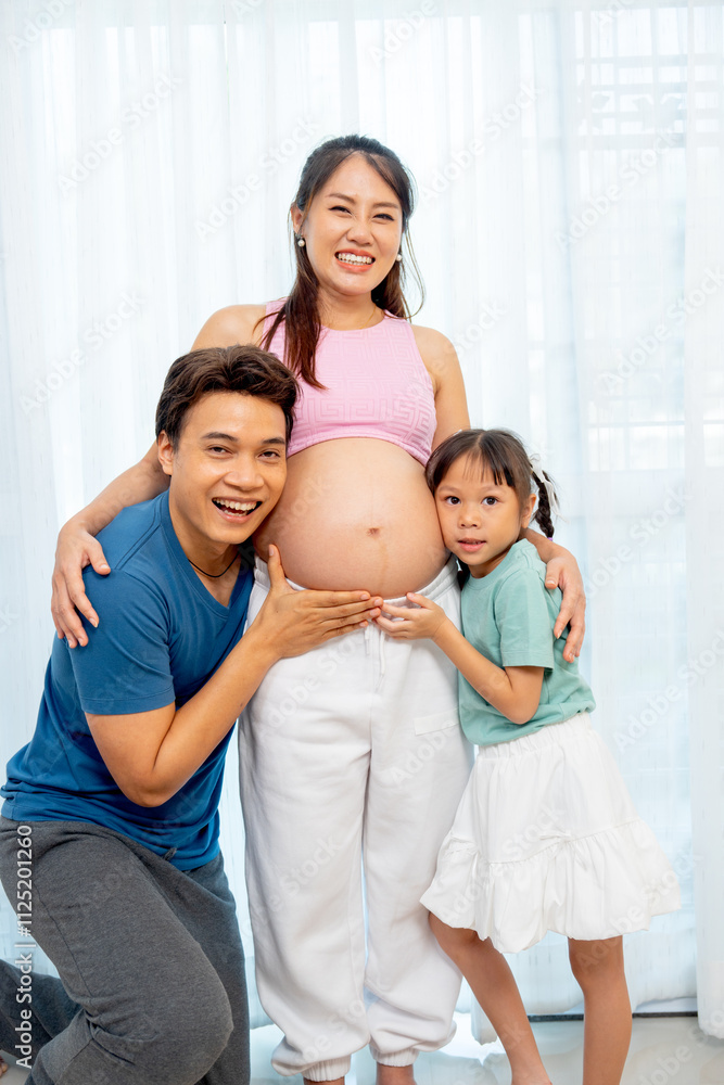 Vertical image of Asian father and little daughter hug and touch to belly of pregnant mother and express happiness with smiling in front of white curtain. Vertical image of Asian father and little daughter hug and touch to belly of pregnant mother and express happiness with smiling in front of white curtain.