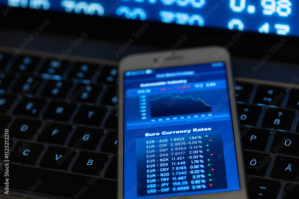 Smartphone with currency exchange rates like Euro, US dollar, British pound, Swiss franc or Japanes yen lays on a laptop. Screen with data in the background. Business, trading, information. Smartphone with currency exchange rates like Euro, US dollar, British pound, Swiss franc or Japanes yen lays on a laptop. Screen with data in the background. Business, trading, information.