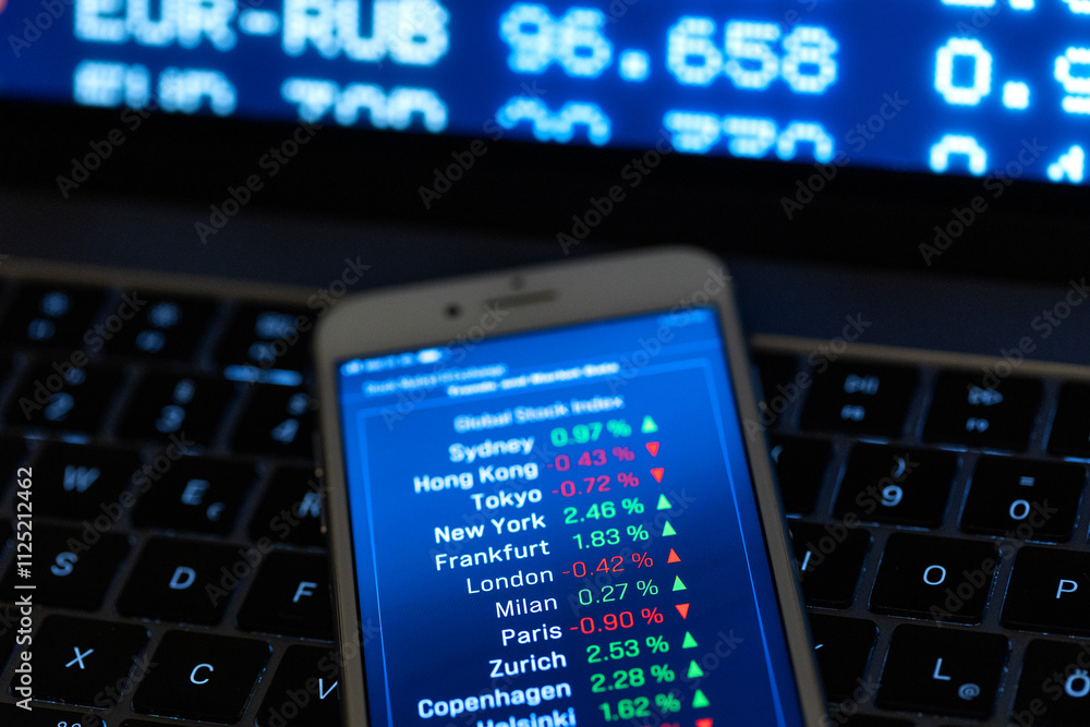 The smartphone is placed on the keyboard of a laptop and displays global stock indices and their changes. Trading, investment, volatility, share price, financial markets, business. The smartphone is placed on the keyboard of a laptop and displays global stock indices and their changes. Trading, investment, volatility, share price, financial markets, business.
