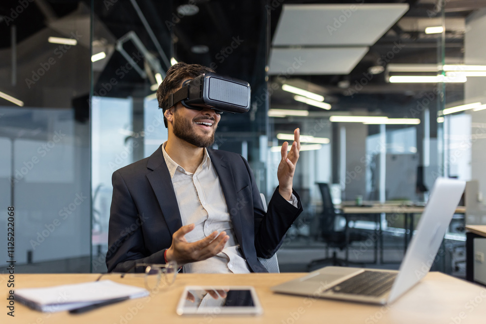 Fototapeta premium A businessman in a modern office uses virtual reality for an immersive experience, showcasing the integration of technology in business environments. A laptop and smartphone are on the desk.