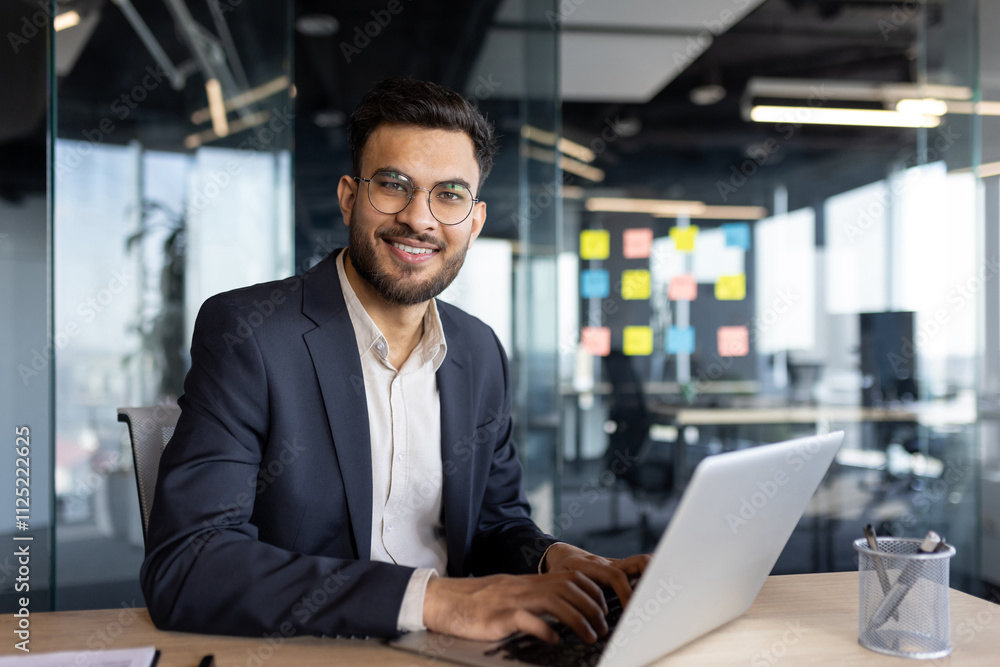 A confident businessman in a suit is working on a laptop in a bright, modern office. The office features glass walls and colorful sticky notes, creating an open and collaborative work environment. A confident businessman in a suit is working on a laptop in a bright, modern office. The office features glass walls and colorful sticky notes, creating an open and collaborative work environment.