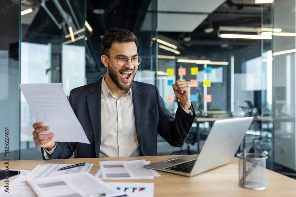 A cheerful businessman in a suit celebrates a successful achievement at his office desk. The image captures a moment of joy and motivation while reviewing documents on a laptop. A cheerful businessman in a suit celebrates a successful achievement at his office desk. The image captures a moment of joy and motivation while reviewing documents on a laptop.