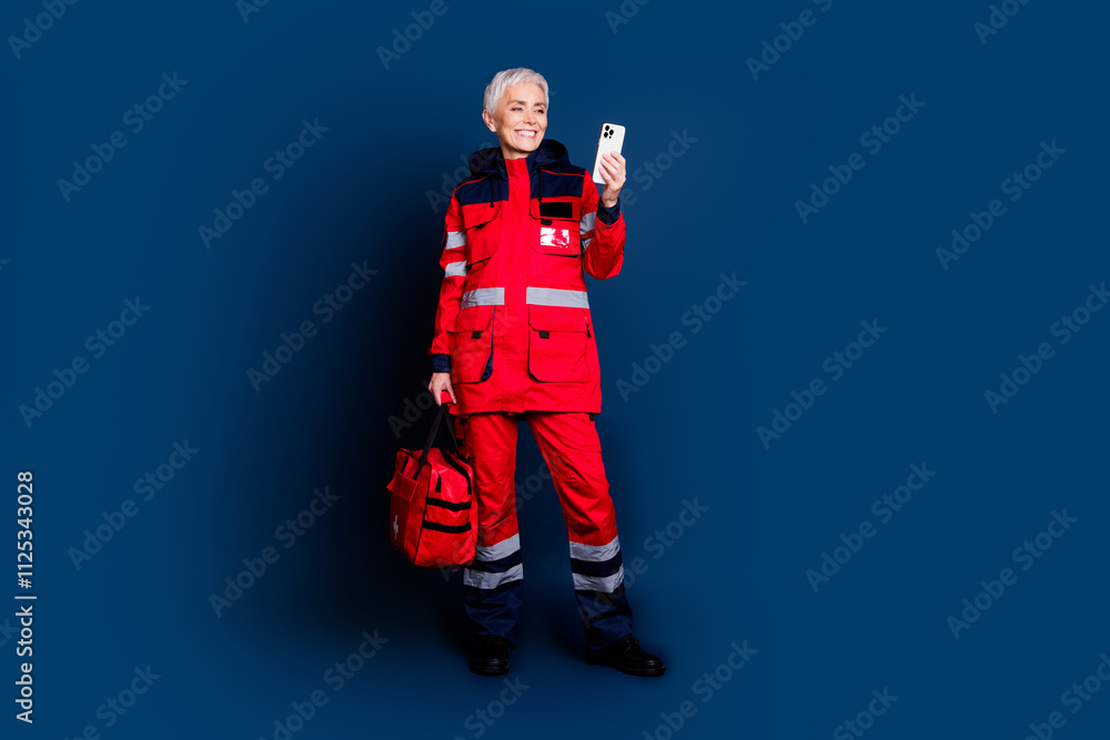 Full length photo of pensioner lady provide emergency medical help hold device dressed red uniform isolated on dark blue color background Full length photo of pensioner lady provide emergency medical help hold device dressed red uniform isolated on dark blue color background