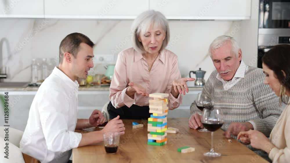 Cheerful senior woman spending free time with husband and adult children at laid-back family home get-together with drinks, concentrating on removing block from tower while playing jenga Cheerful senior woman spending free time with husband and adult children at laid-back family home get-together with drinks, concentrating on removing block from tower while playing jenga