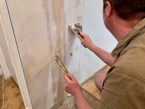 A man uses a putty knife and plaster to smooth a wall seam on plasterboard, preparing the surface for painting during a DIY home renovation project.