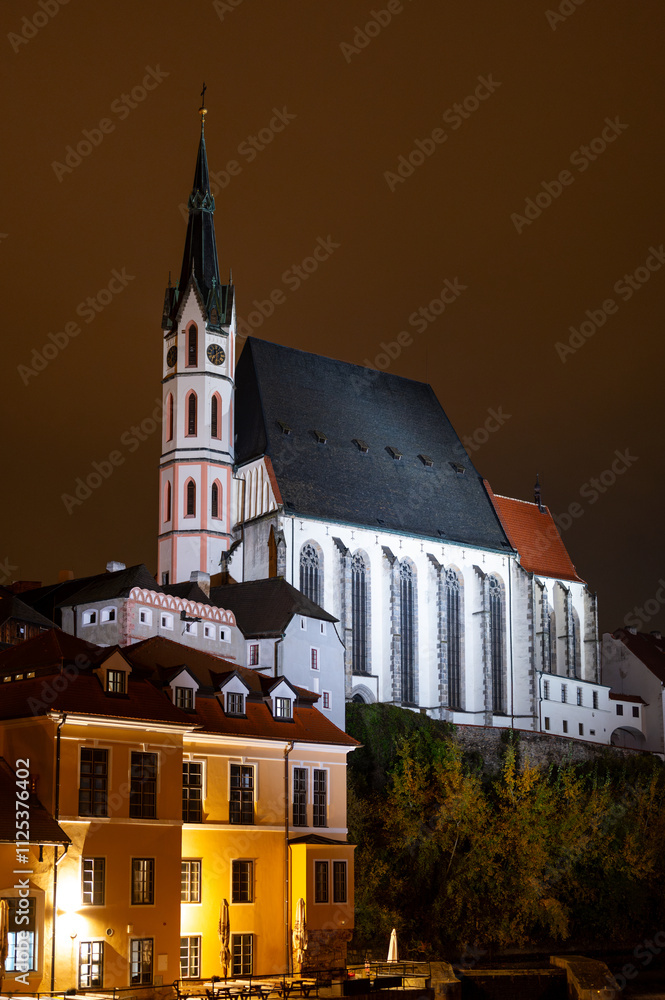 A night view of the Church of St. Vitus in Cesky Krumlov, beautifully illuminated by lights, standing tall against the dark sky, highlighting its architectural details