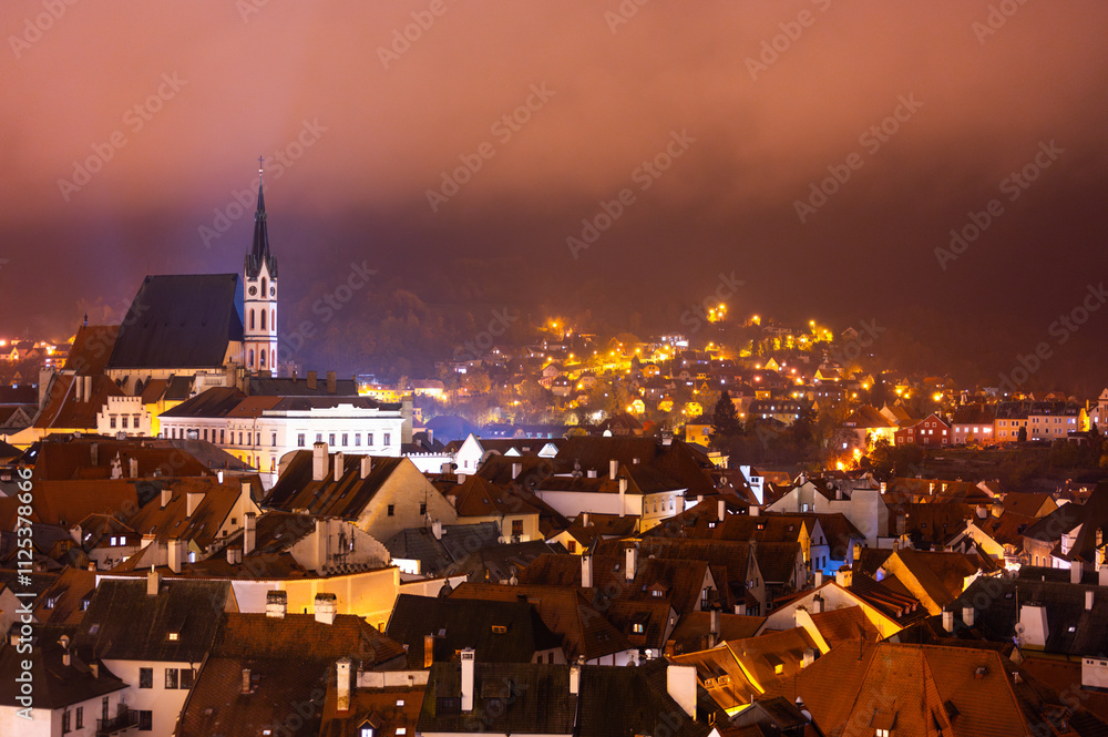 A panoramic night view of Cesky Krumlov, showcasing the rooftops, illuminated buildings, and the Church of St. Vitus beautifully lit up against the dark sky
