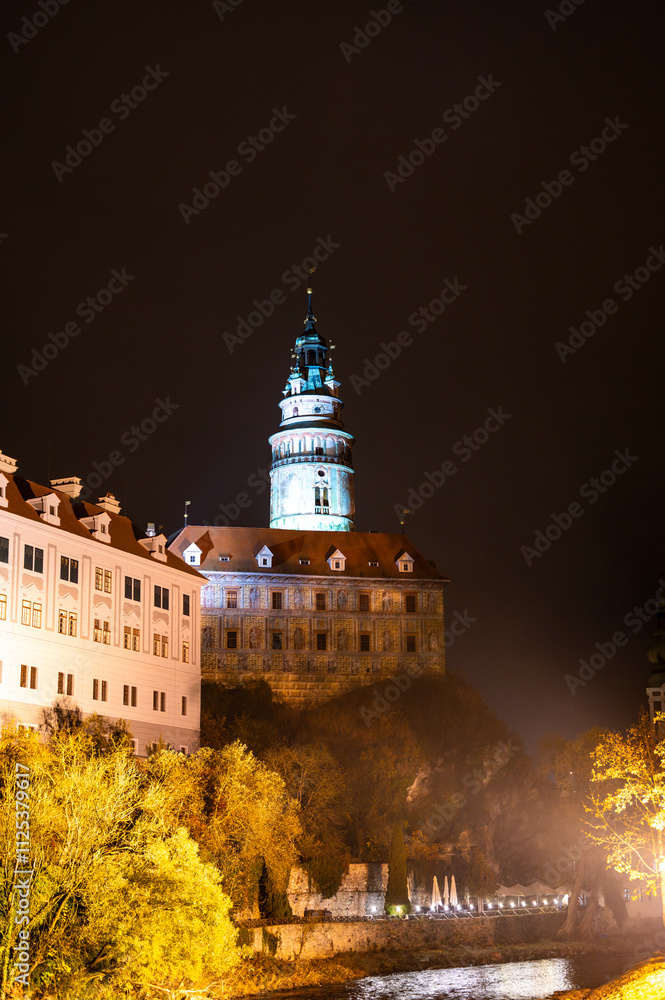 A stunning night view of the illuminated tower of Cesky Krumlov Castle towering above the Vltava River, creating a magical atmosphere with the reflection in the water