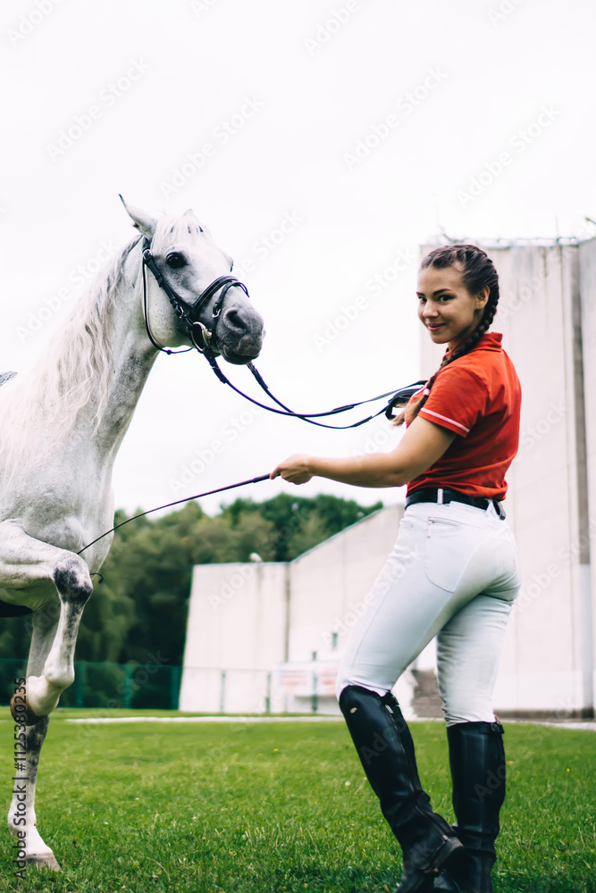 Obraz premium Full length portrait of Caucasian jockey girl posing during dressage competition at nature, female horseback rider looking at camera while white champion stallion animal showing stunt outdoors