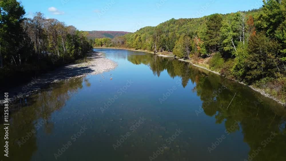 Drone video rising above a clear river which flows into an autumn-colored valley surrounded by a road and agricultural fields. Drone video rising above a clear river which flows into an autumn-colored valley surrounded by a road and agricultural fields.