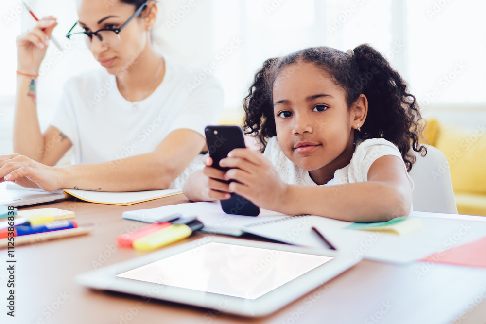 Mother using laptop and daughter with phone at table in living room Mother using laptop and daughter with phone at table in living room