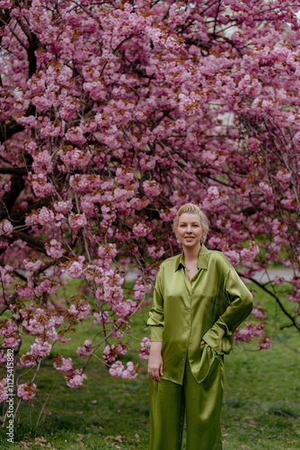 A blonde woman in green outfit poses surrounded by cherry blossoms outdoors