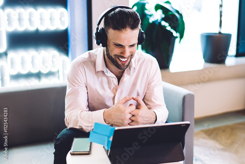 Happy Caucasian man watching positive video via portable touch pad, successful male blogger in electronic headphones for noise cancellation searching funny publication for reading on leisure photo