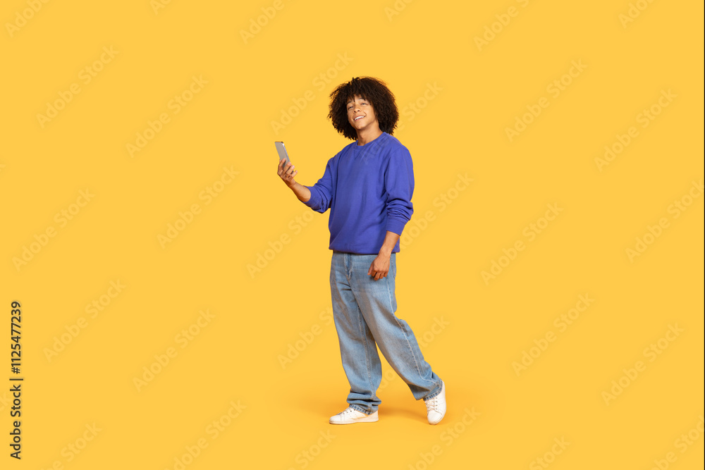 A young black man smiles while standing in relaxed jeans and a sweater, confidently engaging with his smartphone. The vibrant yellow background creates a cheerful atmosphere. A young black man smiles while standing in relaxed jeans and a sweater, confidently engaging with his smartphone. The vibrant yellow background creates a cheerful atmosphere.