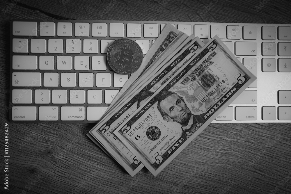 A Bitcoin coin lies on the desk next to a few dollar notes on a computer keyboard