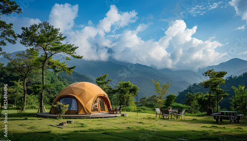 big brown dome camping tent in a green grass with trees and mountains. Taken in an afternoon with a clear blue sky with white clouds. There're chairs, table and camping equipments nearby