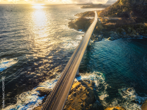 Aerial view of Djupfjord bridge crossing the fjord with snowy mountains, colorful sky in golden hour in winter. Moskenes, Lofoten islands, Norway. Top view of road, snowy rocks, blue sea at sunset photo