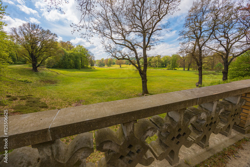 Beautiful landscape of big green park with green glades, trees at cloudy sunny morning seen from small bridge with concrete fence of in park