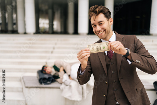 Cheerful successful businessman in suit smiling looking at camera as holding dollar bill while homeless person lying on steps of urban building, showing social and economic disparities. photo