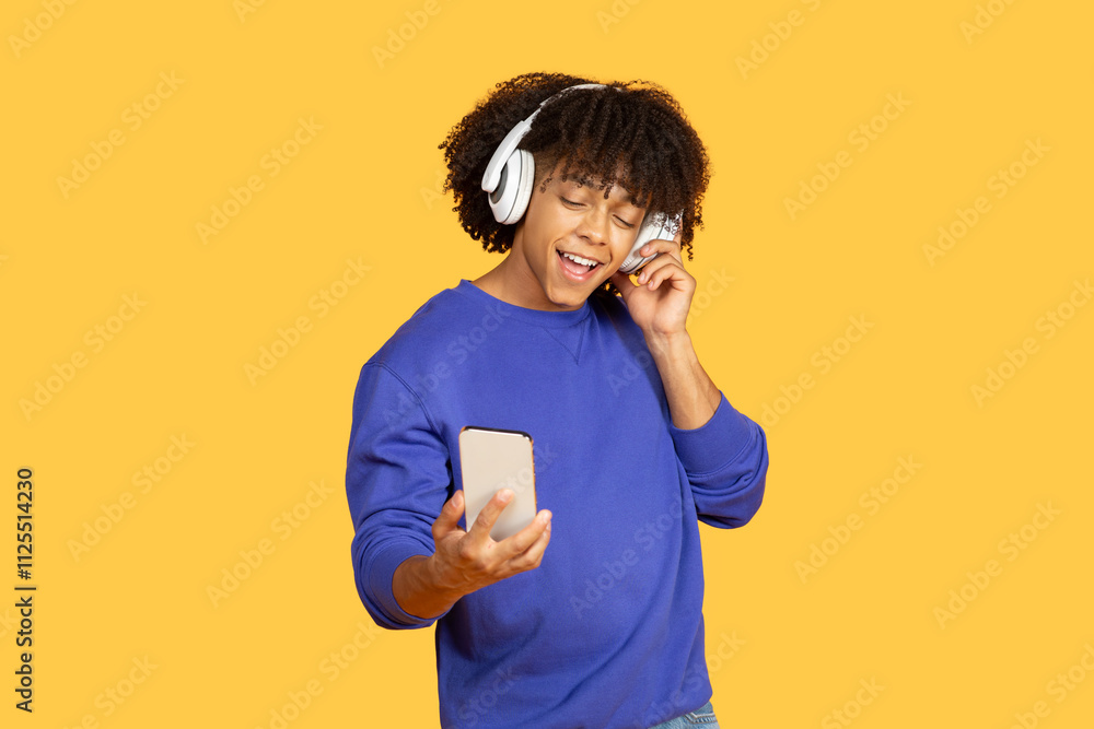A young Black man with curly hair wearing white headphones smiles while listening to music. He holds a smartphone and is immersed in the upbeat rhythm against a bright yellow backdrop. A young Black man with curly hair wearing white headphones smiles while listening to music. He holds a smartphone and is immersed in the upbeat rhythm against a bright yellow backdrop.