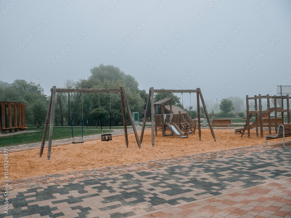Fototapeta premium Modern playground on a sand surface and built with use wooden organic materials. No people, fog in the background. Concrete footpath around the play ground area.