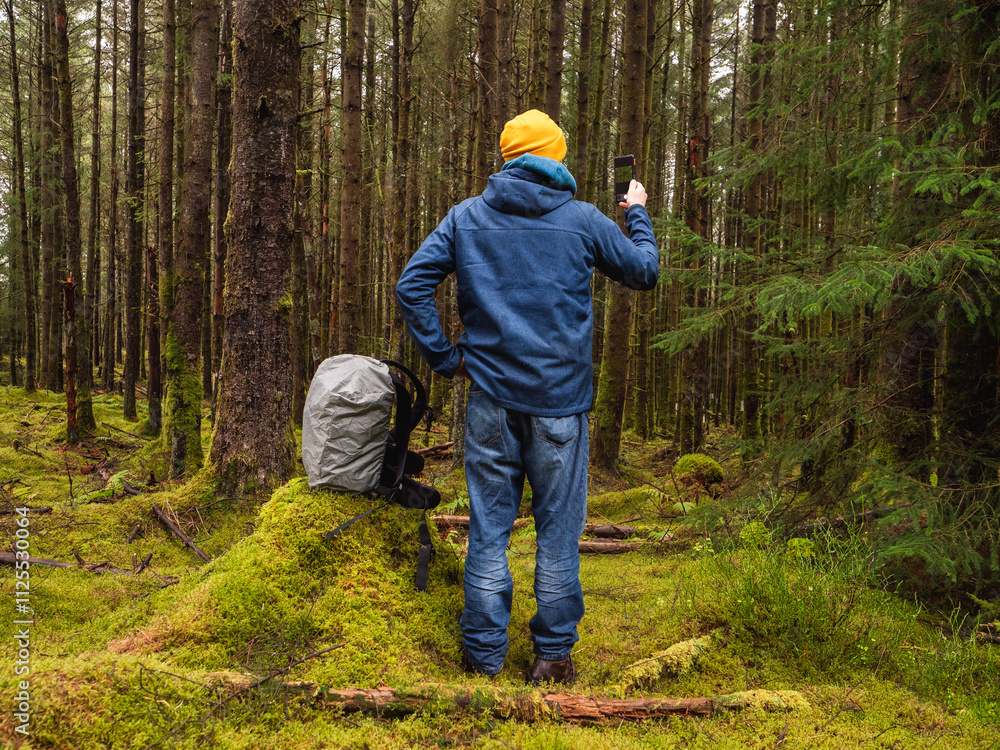 Male tourist taking pictures on phone, his backpack on a tree stump. Scene is peaceful and serene, as the man is surrounded by nature. Travel and tourism theme. Model in blue jacket and yellow hat. Male tourist taking pictures on phone, his backpack on a tree stump. Scene is peaceful and serene, as the man is surrounded by nature. Travel and tourism theme. Model in blue jacket and yellow hat.