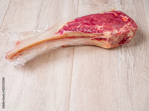 Fine tomahawk steak in a plastic wrap on a wooden table. Premium crat red meat on the bone. High value product with fantastic taste. Farming and agriculture industry. Preserving freshness photo