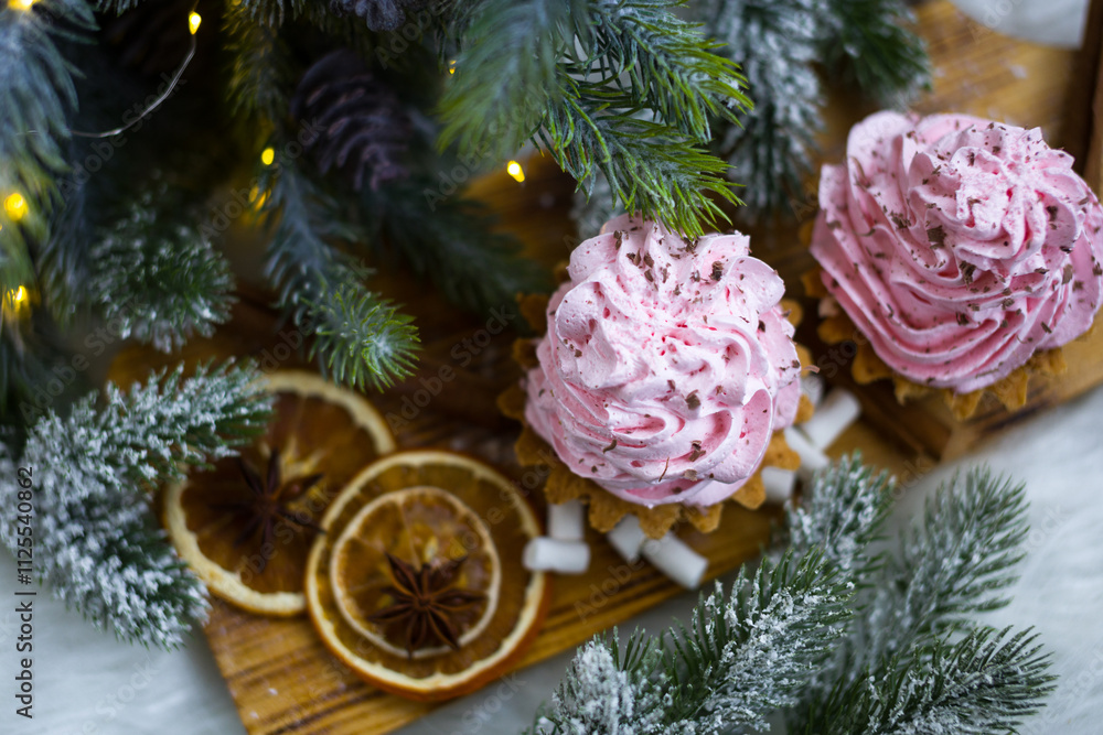 Pink cakes on a wooden board under a Christmas tree with lights. A cup with marshmallows and orange slices Pink cakes on a wooden board under a Christmas tree with lights. A cup with marshmallows and orange slices