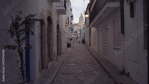 A quiet narrow street in Cadaques during sunset, with a glowing streetlight and the church visible in the background. A parked motorbike is seen on the sidewalk, capturing the peaceful atmosphere photo