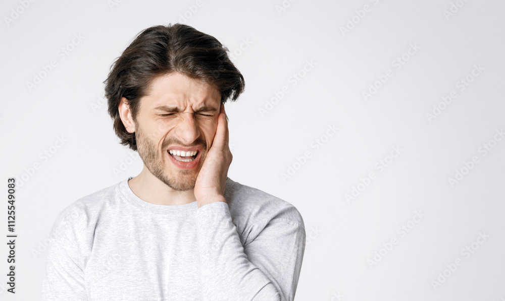 Sharp pain in ears. Man grimaces and holds hand to sore spot isolated on gray background, studio shot Sharp pain in ears. Man grimaces and holds hand to sore spot isolated on gray background, studio shot