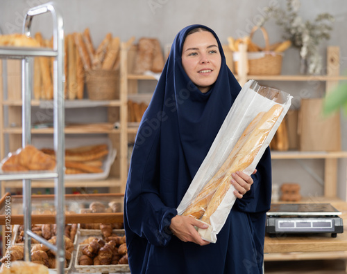 Positive interested young female customer of European features, wearing traditional Muslim blue abaya holding package of fresh French baguettes while shopping in cozy local bakery..
