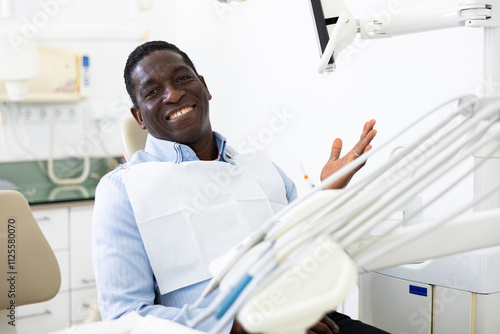 Portrait of smiling african-american man sitting on dental chair and making palm gesture.. photo