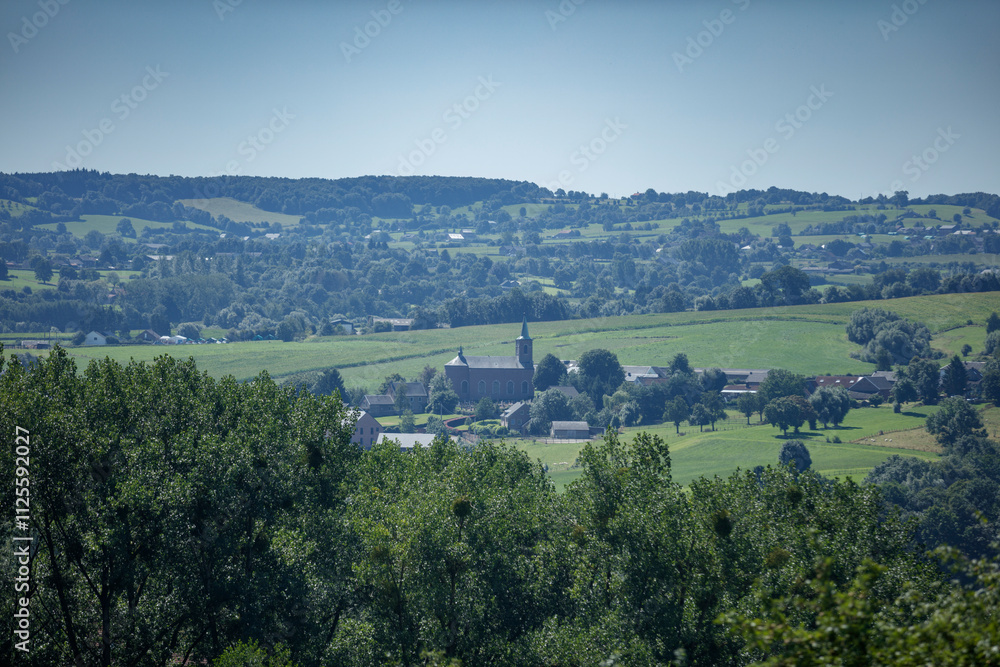 Naklejka premium Panoramic view of a rural European village nestled in a valley, with fields, trees, and a church steeple visible. Tranquil countryside scene.