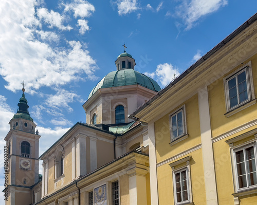 Ljubljana, Slovenia - June 28, 2024: South facade of Saint Nicholas's Cathedral with clock tower seen from Ciril-Metodov shopping street under blue cloudscape.  photo