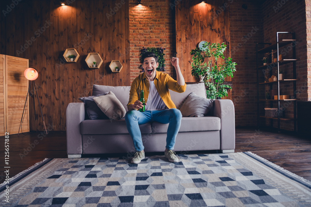 Obraz premium Excited young man in stylish shirt enjoys leisure time at home, holding a drink, celebrating in a comfortable and modern living
