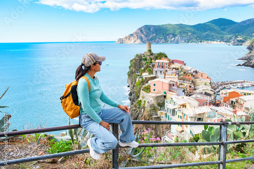 A woman traveler sits on a railing, gazing at the stunning coastline of Cinque Terre. The vibrant houses perched on the cliffs and the sparkling sea create a picturesque backdrop, Vernazza Italy