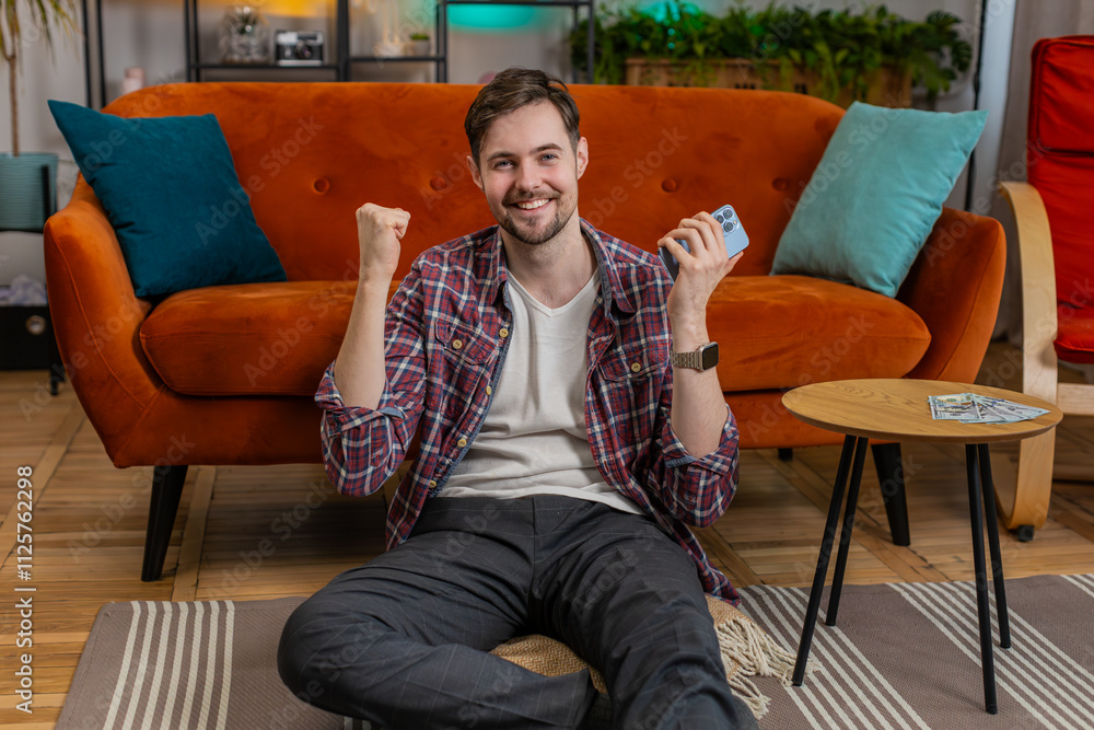 Fototapeta premium Amazed excited young man winner holds smartphone reading good news amazed by online bet bid game lottery jackpot casino win. Happy Caucasian guy overjoyed by victory success shows thumbs up at home