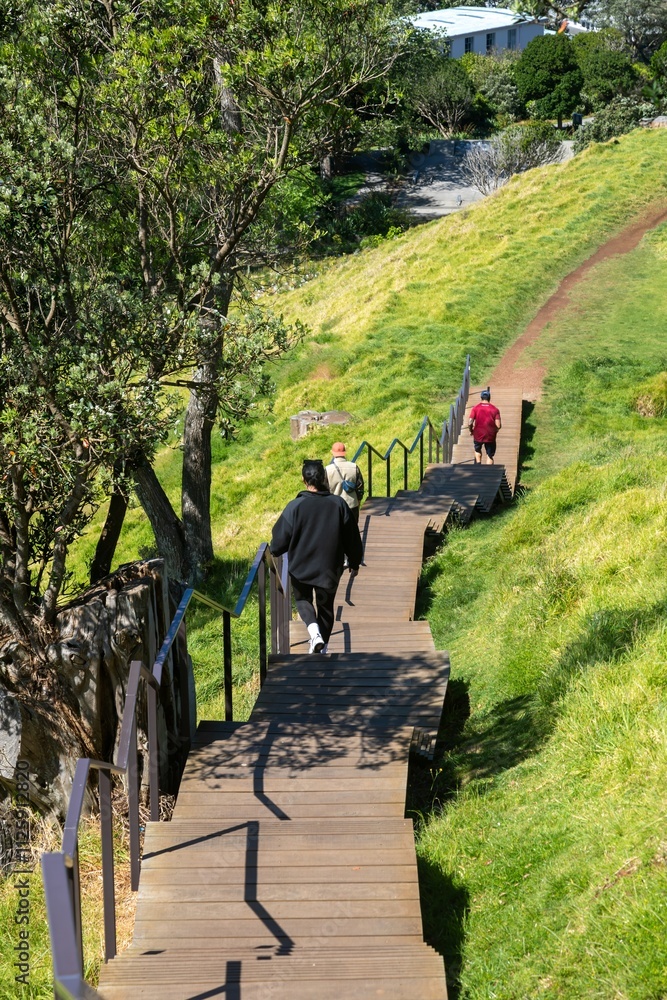 MANGERE MOUNTAIN, AUCKLAND, AUCKLAND, NEW ZEALAND, Hikers ascend a wooden staircase through on the grassy volcanic hilltop park, overlooking a city and harbor. MANGERE MOUNTAIN, AUCKLAND, AUCKLAND, NEW ZEALAND, Hikers ascend a wooden staircase through on the grassy volcanic hilltop park, overlooking a city and harbor.