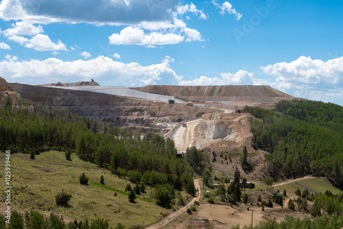 Industrial open pit gold mining site surrounded by green forestry under a blue sky with clouds, showcasing modern extraction techniques in a remote area during daytime