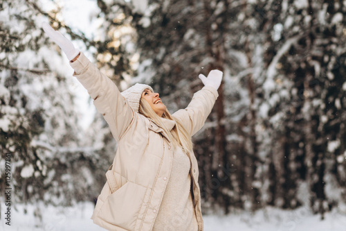 Joyful woman in warm cozy clothes stands among the snowy forest happily raising her hands up, enjoying the winter time.