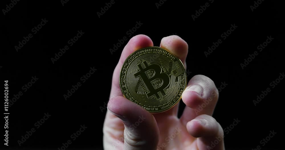 A hand holds a shiny golden bitcoin in sharp focus against a dramatic black background
