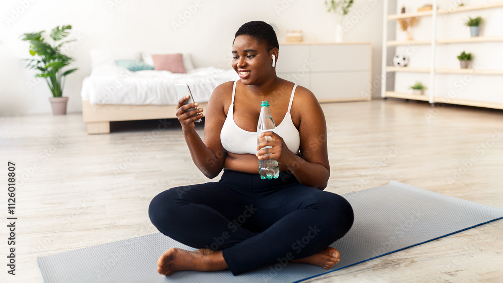 Plus size young black woman with bottle of water and cellphone surfing web on break from home training, checking music online. Curvy African American female using mobile phone after workout Plus size young black woman with bottle of water and cellphone surfing web on break from home training, checking music online. Curvy African American female using mobile phone after workout
