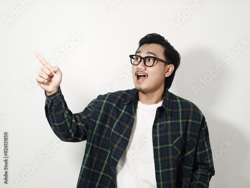 A young Asian man in a plaid shirt looks surprised, pointing enthusiastically at something off-frame against a white background