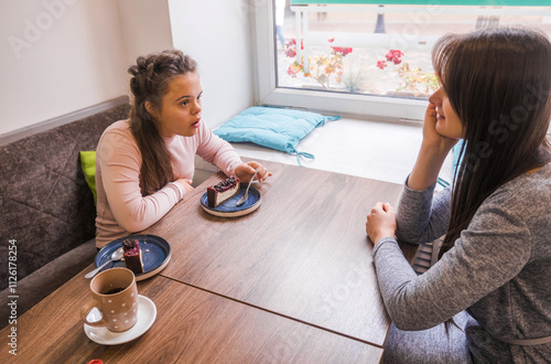 Two young women are enjoying a coffee break together at a cafe. One of the women has Down syndrome; they are engaged in conversation, sharing dessert, and demonstrating a genuine connection. 
