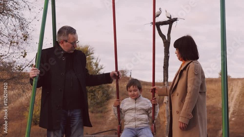 Parents swing son on swings infusing surroundings with joy in park. Parents amid kaleidoscope of colours with son share moments. Parents swinging son on swing on park playground enjoying experience photo