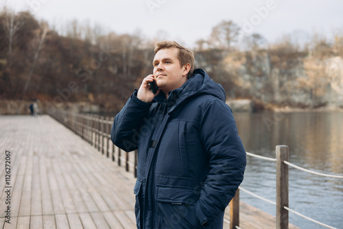 Obraz na płótnie Adult 30s man hiker stand in a natural setting, smile while using his mobile phone outdoors. Male standing on the lake wooden pier. Happy man talk phone, walking near river. Phone Communication w salonie