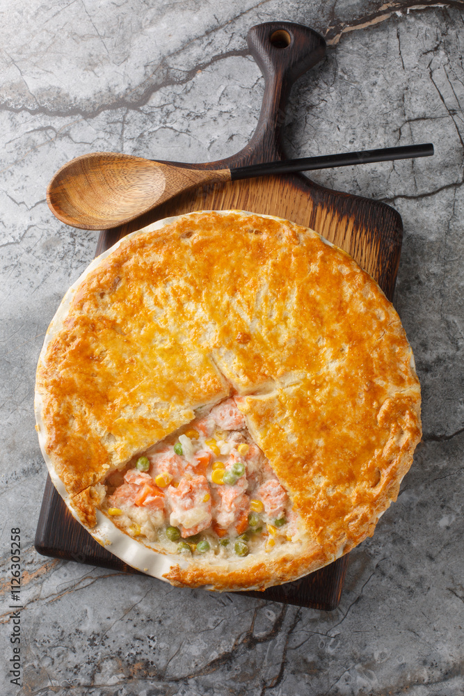 Pot pie with red fish, green peas, carrots, corn and cream sauce covered with crispy dough close-up in a baking dish on the table. Vertical top view from above