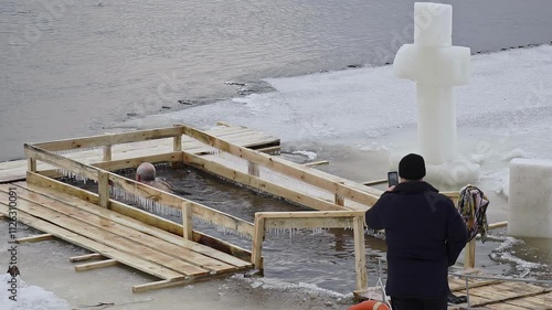 Overweight adult man with naked torso dives 3 times into ice hole of river in winter. Orthodox believer purges sins by diving into water. Unusual religious ritual. Bathing in feast of Epiphany. Faith photo