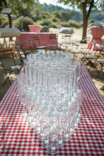 Many clear glasses, stacked, sit on a red and white checkered tablecloth outdoors.  A picnic-style setting. photo