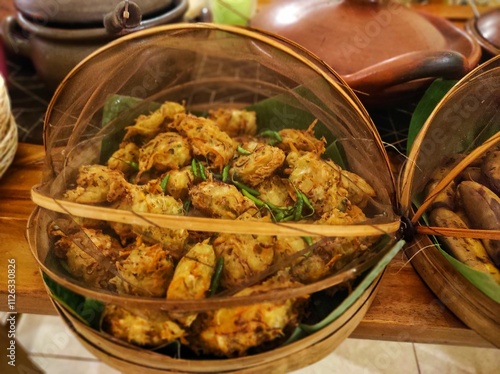 Traditional buffet with boiled bananas and fried fritters on banana leaves, covered with bamboo and cloth. Clay pot in the background adds rustic charm, highlighting Indonesian culinary heritage.